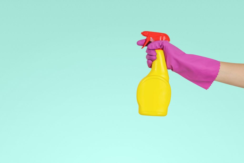 Colorful photo of pink glove-wearing hand holding a yellow spray bottle. Embassy Rabat.