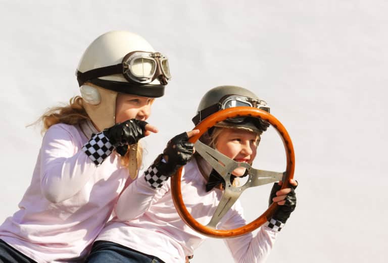 Twin sisters play race car driver sitting in their imaginative sports car.
They play happily wearing vintage helmets, goggles and use an
old-timer steering wheel they found in the garage.